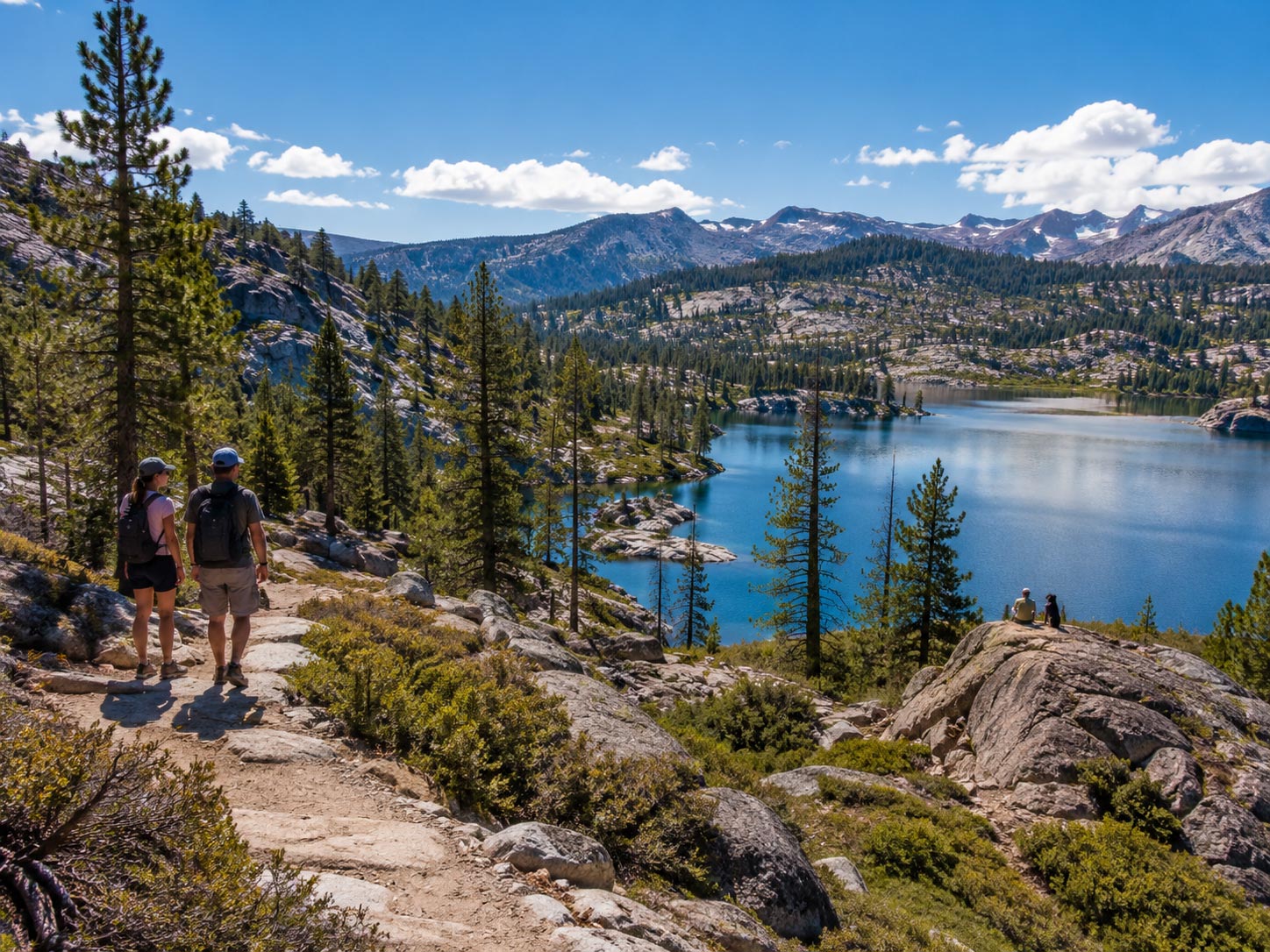 Two hikers walking along a rocky mountain trail overlooking a bright blue alpine lake surrounded by pine trees and granite peaks.